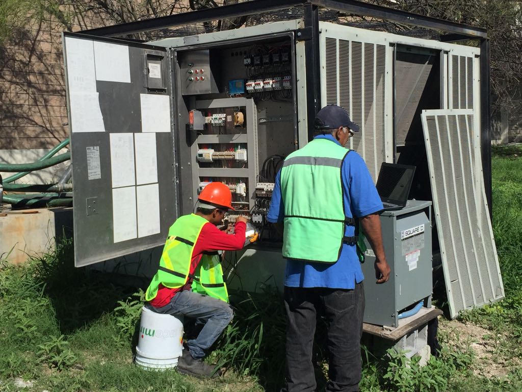 Dos técnicos con chalecos reflectantes y cascos revisan un panel eléctrico exterior abierto, con una computadora portátil sobre una unidad Square D cercana.